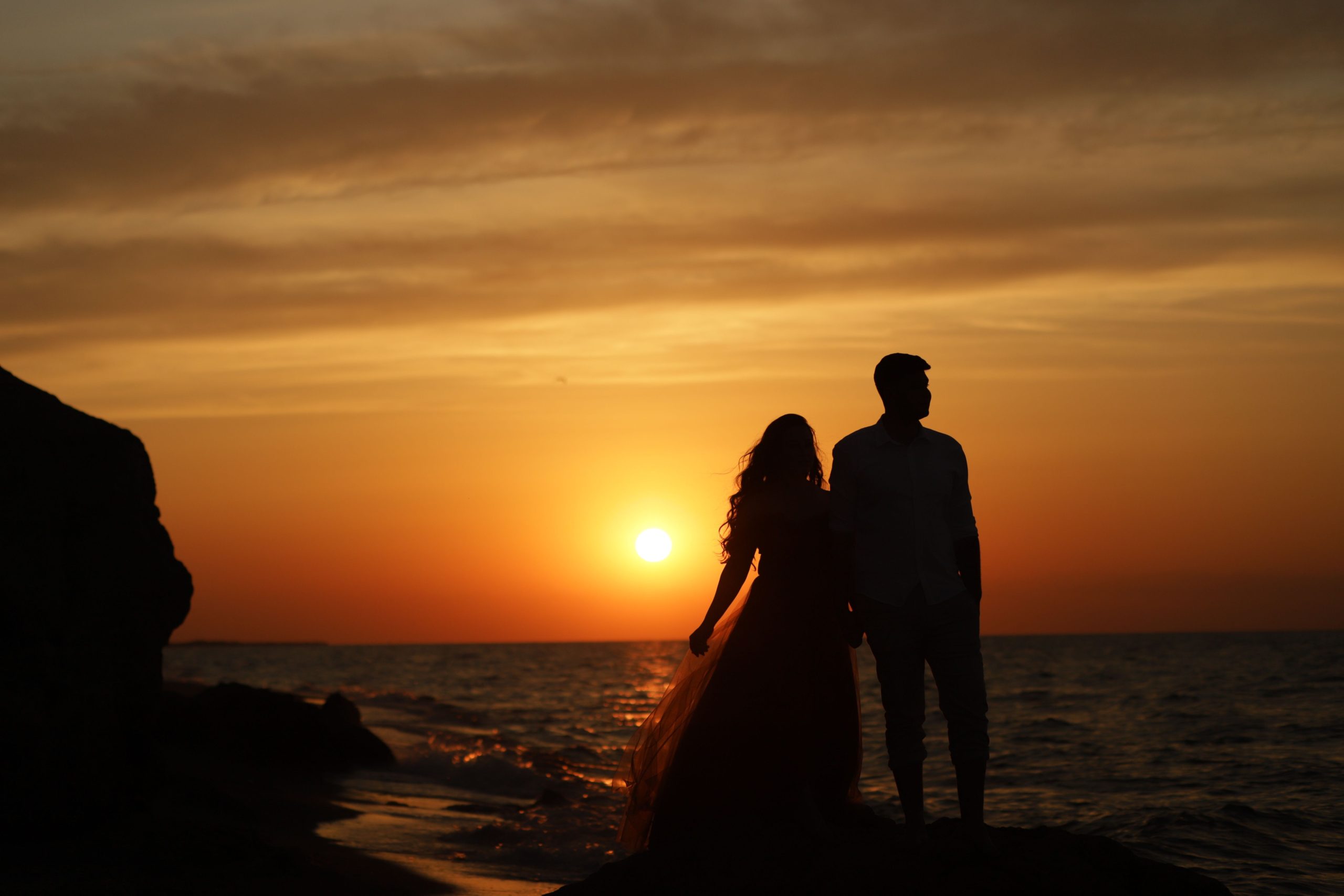 Silhouette of a couple holding hands by the sea at sunset, creating a romantic and serene mood.